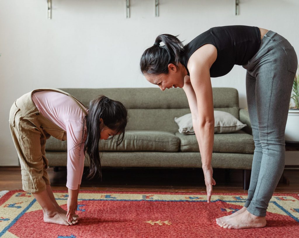 a mother wearing a black top and grey yoga pants and a young daughter wearing a long sleeved pink shirt and brown overalls are trying to touch their feet.