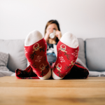 a woman is sitting on a grey couch and drinking from a coffee mug. She has her feet on the wooden coffee table and is wearing decorative reindeer socks.