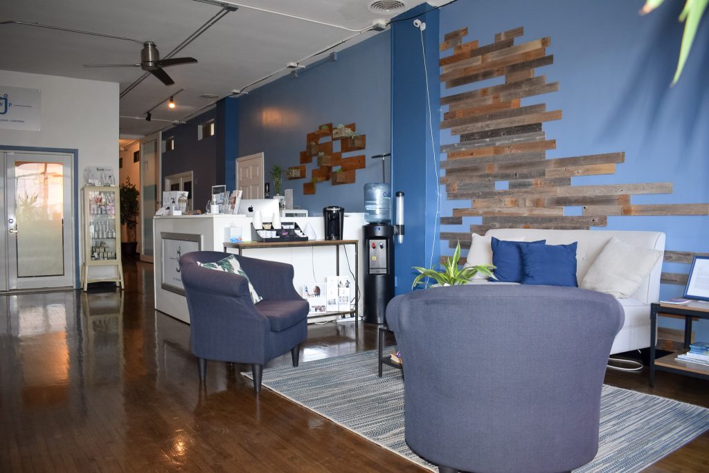 the inside lobby of JWP and EA. It has a blue wall, brown wooden floors, two single navy blue couches, a white loveseat couch, a white receptionist desk, and a water cooler.