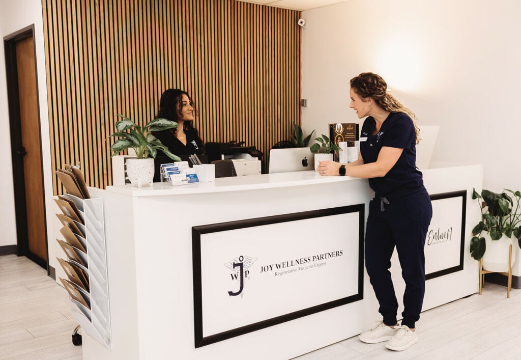 Joy Wellness Partners' founder Carol stands, smiling cheerfully, conversing with front desk receptionist.